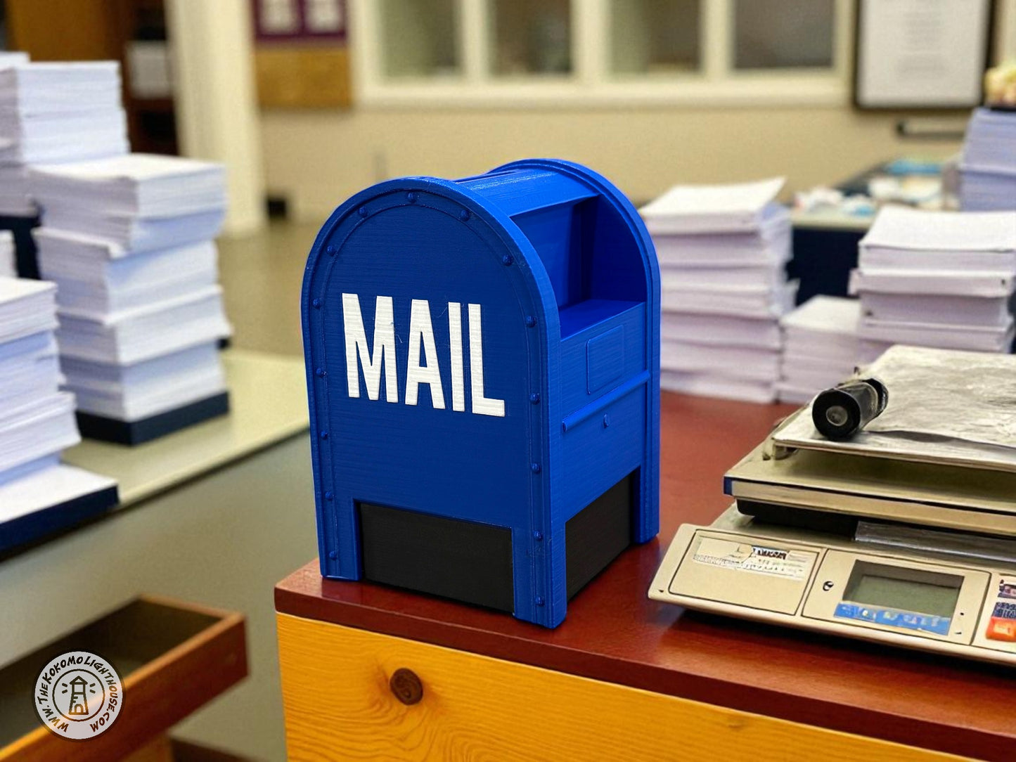 Blue piggy bank mailbox labeled 'MAIL' on a desk with stacks of papers and a scale in an office setting.