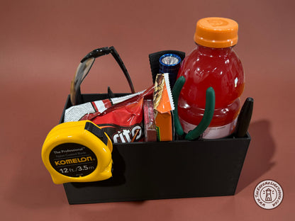 A rectangular black fork truck caddy holding a gatorade, safety glasses, a tape measure, and other misc supplies and snacks with a maroon background.