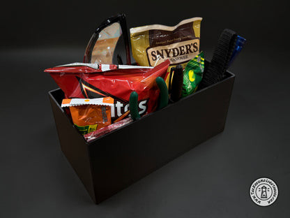 A rectangular black fork truck caddy holding a bag of chips, safety glasses, a tape measure, and other misc supplies and snacks with a dark background.