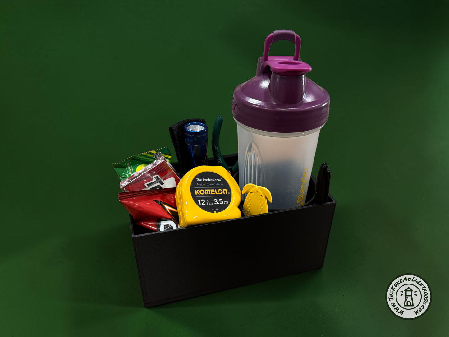 A rectangular black fork truck caddy holding a shaker bottle, safety glasses, a tape measure, and other misc supplies and snacks with a dark green background.