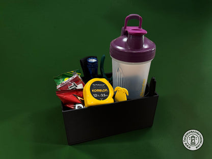 A rectangular black fork truck caddy holding a shaker bottle, safety glasses, a tape measure, and other misc supplies and snacks with a dark green background.