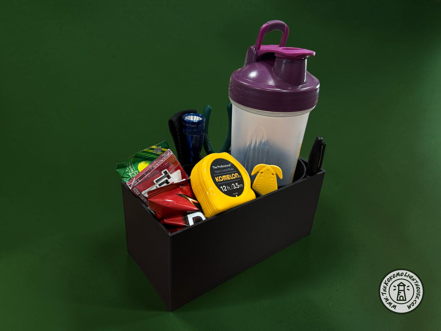 A rectangular black fork truck caddy holding a shaker bottle, safety glasses, a tape measure, and other misc supplies and snacks with a dark green background.