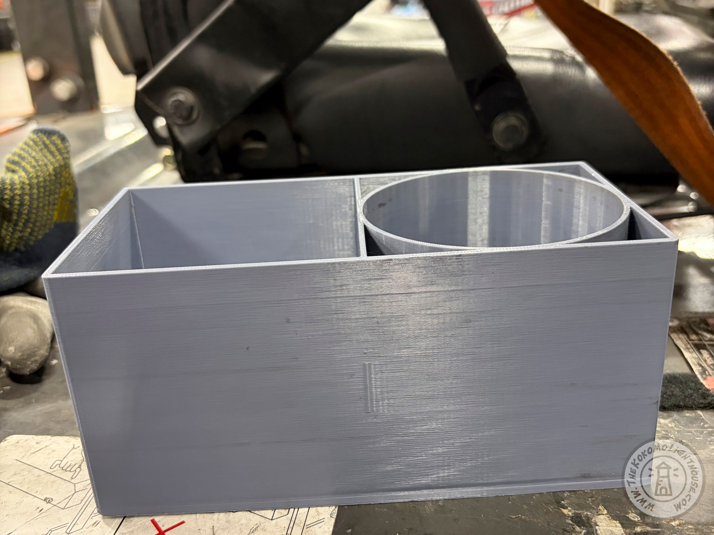 A light gray 3D-printed dual-compartment organizer tray with a rectangular main section and a cylindrical cup-like compartment, shown on a forklift.