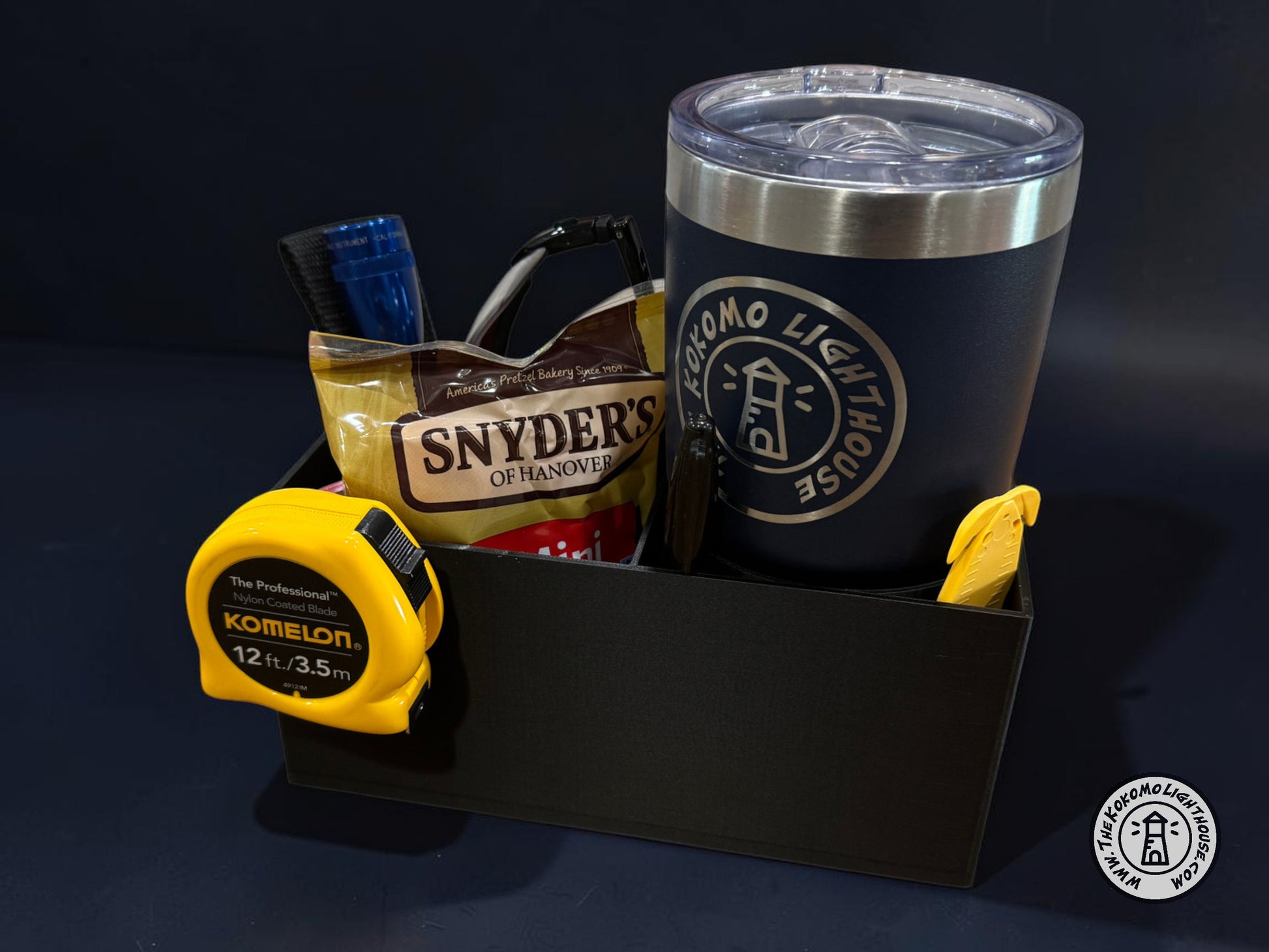 A rectangular black fork truck caddy holding a tumbler, safety glasses, a tape measure, and other misc supplies and snacks with a dark blue background.