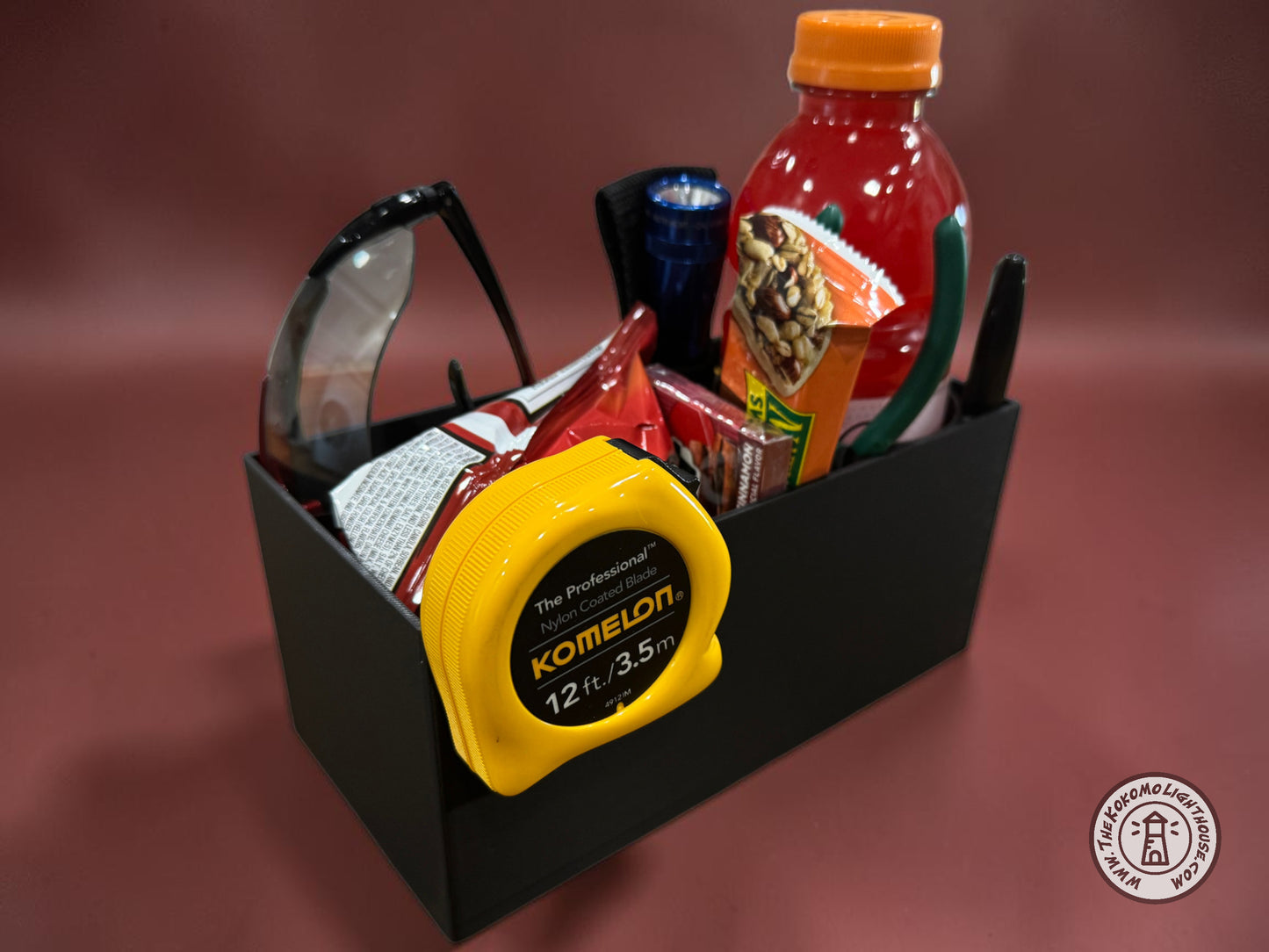 A rectangular black fork truck caddy holding a gatorade, safety glasses, a tape measure, and other misc supplies and snacks with a maroon background.