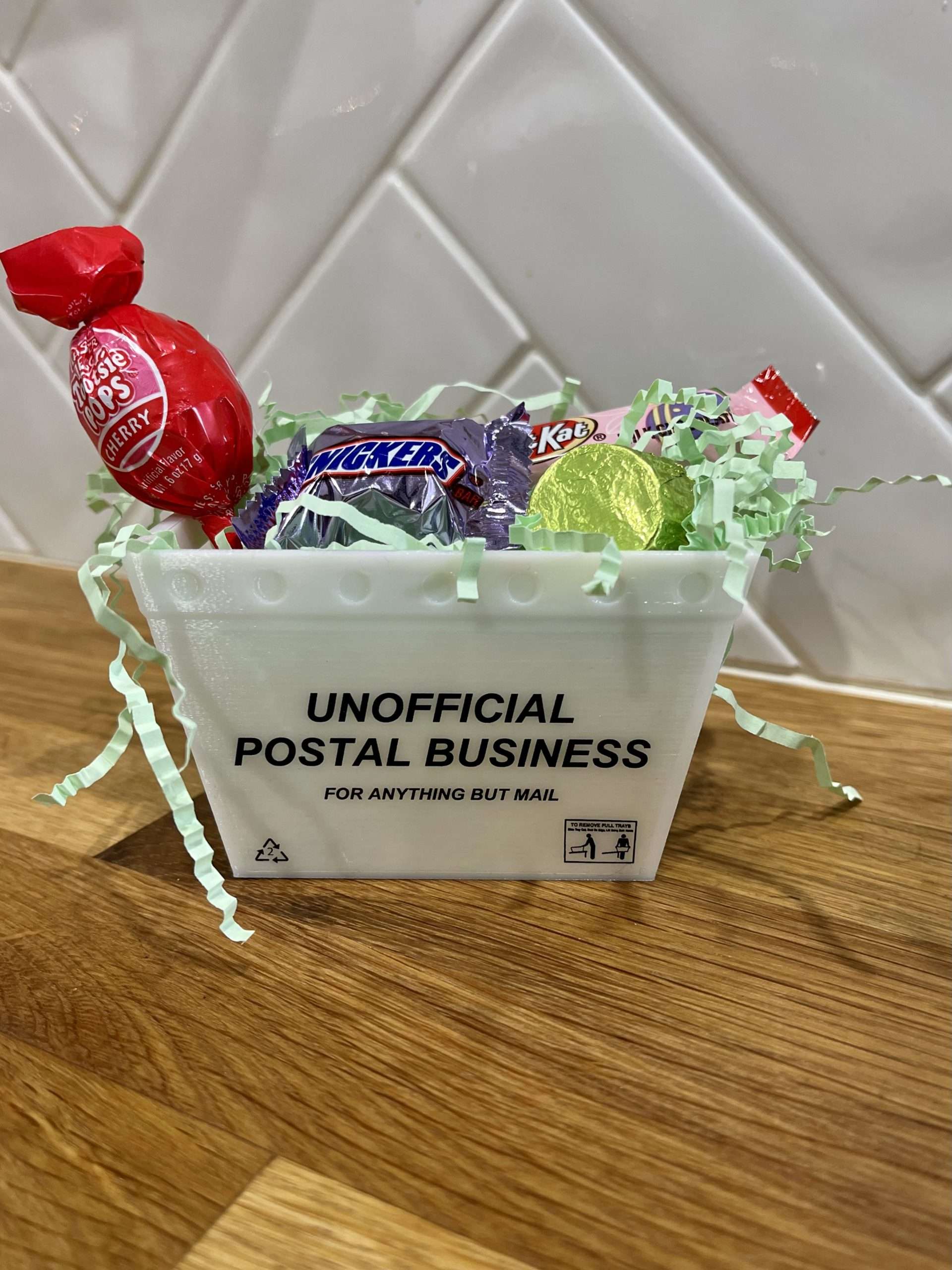 A mini mail tote on a wooden counter with a white tile background filled with easter grass and various candies