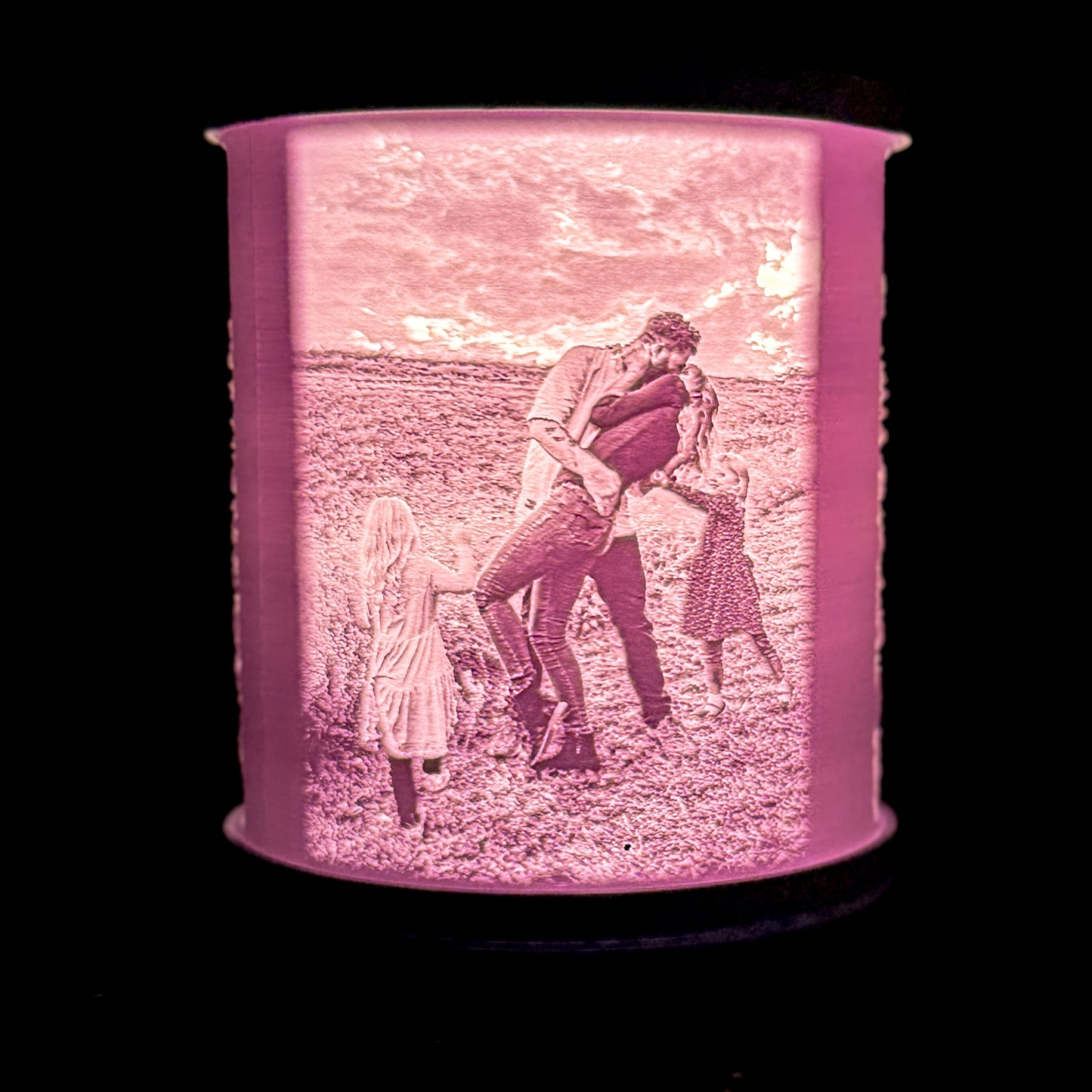 Lit family photo lamp showing adults kissing and children pulling them apart in an outdoor field, with the photo details glowing through the white shade.