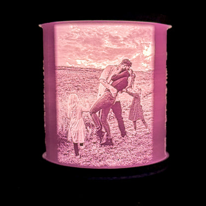 Lit family photo lamp showing adults kissing and children pulling them apart in an outdoor field, with the photo details glowing through the white shade.