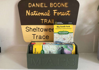 A large green map storage box with 'MAPS' embossed on the front, containing various folded maps. The box is placed on a shelf with a sign reading 'DANIEL BOONE NATIONAL FOREST TRAIL - Sheltowee Trace'.