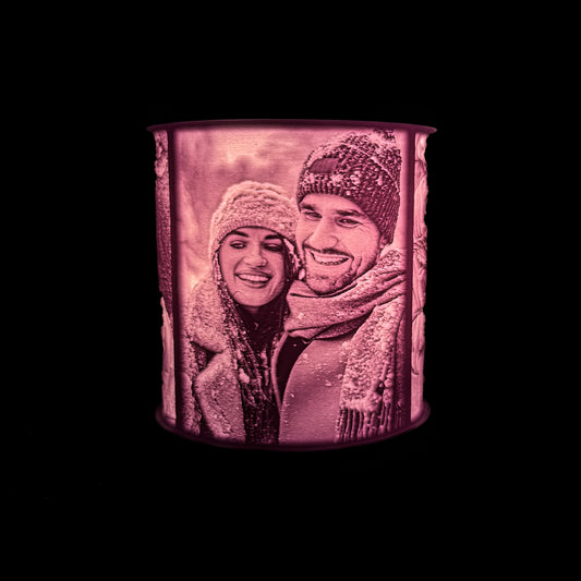 Lit cylindrical lithophane photo lamp showing a smiling couple in winter clothing on anniversary vacation, with the photo glowing through the white 3D printed shade with a dark background.