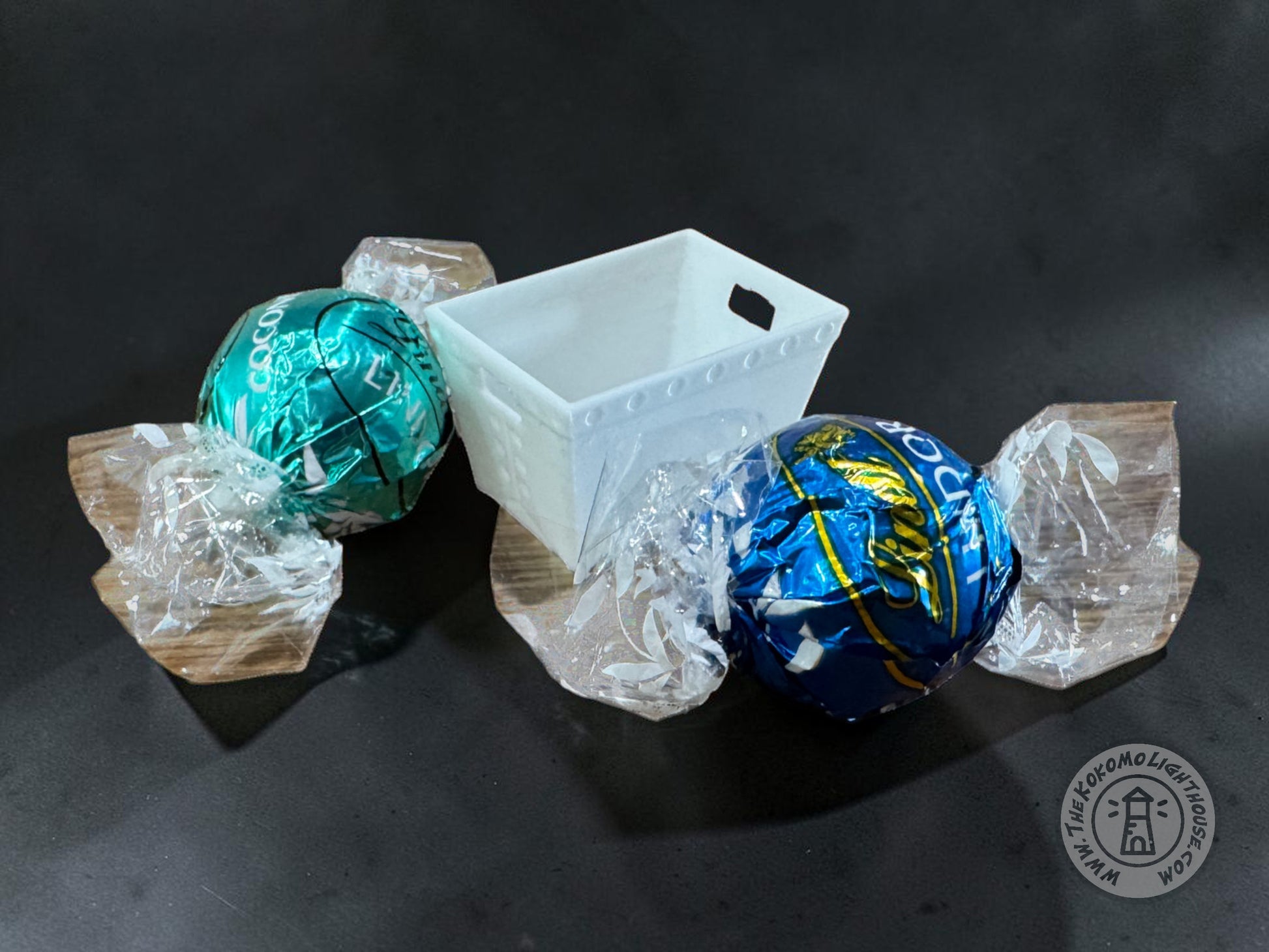 Two foil-wrapped round candies, one teal-green and one blue with yellow graphics, arranged around a small white postal dps tub on a dark background and a circular watermark appears in the lower right corner.