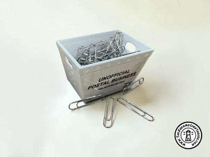 a miniature postal tub filled with paper clips, including some outside the tub on a light background